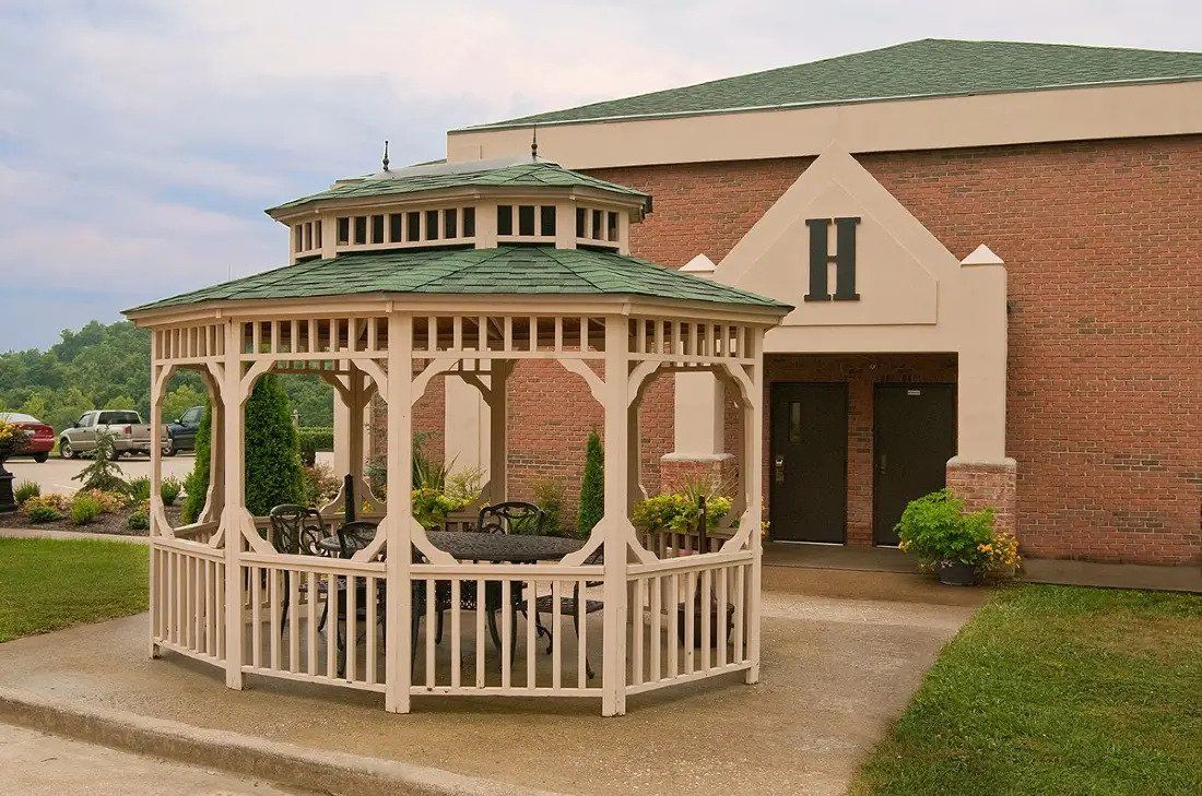 A white wooden gazebo near a building with a green roof and large window.